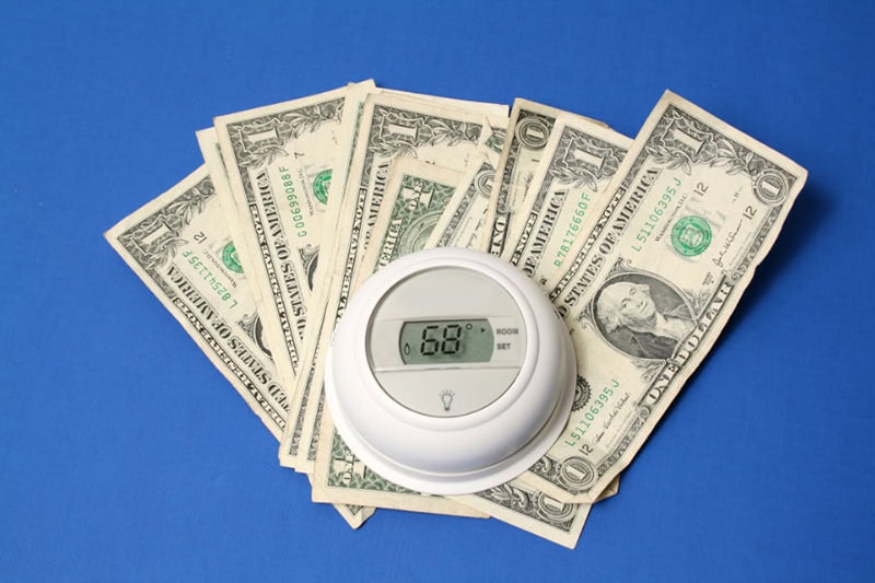 Spring Money-Saving Tips. Photo of a thermostat on top of a pile of cash on a blue table or surface.