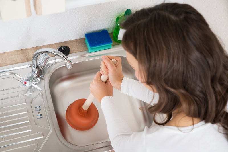 Why Do My Drains Keep Clogging? Happy Woman Using Plunger In Kitchen Sink At Home.